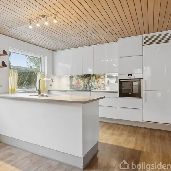 Hvidt køkken med højglansskabe og indbygget ovn. Køkkenø med vask. Loftet er træbeklædt, og gulvet er trægulv. Vindue ud mod haven, åbne hylder på væggen.