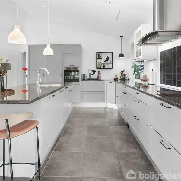 Et lyst, moderne køkken med en L-formet køkkenø, sorte bordplader, grå skuffer og skabe. To barstole står ved øen. Baggrunden har hvide vægge, hængende lamper og planter.