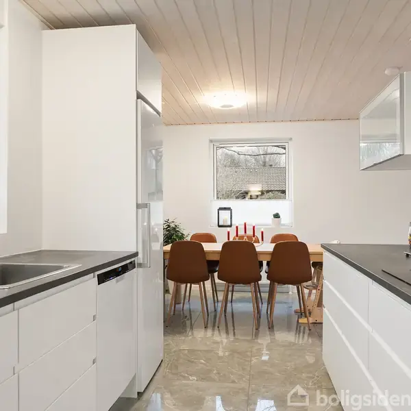 Et moderne køkken med hvide skabe indeholder en stålvask og opvaskemaskine. I baggrunden er et spisebord med seks brune stole, placeret nær et vindue med udsigt til en frodig baghave.