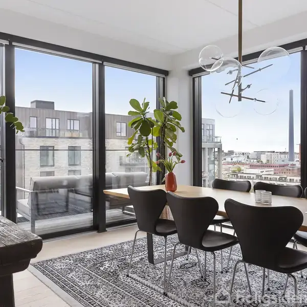 Spisebord med sorte stole står i et moderne rum med store vinduer. Udsigten viser bylandskab og fabriksbygninger. En dekorativ lampe hænger over bordet, og en plante står ved siden af.