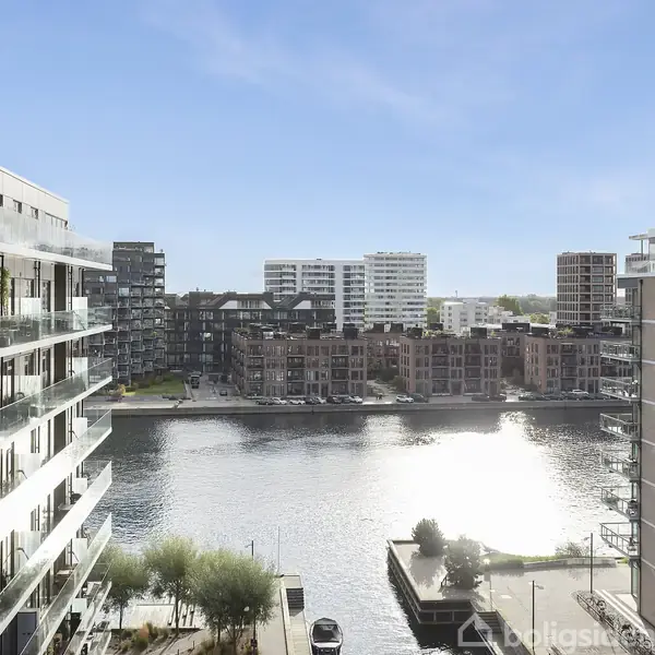Floder mellem moderne boligbyggerier, solen spejler sig i vandet. Omkringliggende lejlighedskomplekser har store altaner. I baggrunden ses flere højhuse under en klar blå himmel.