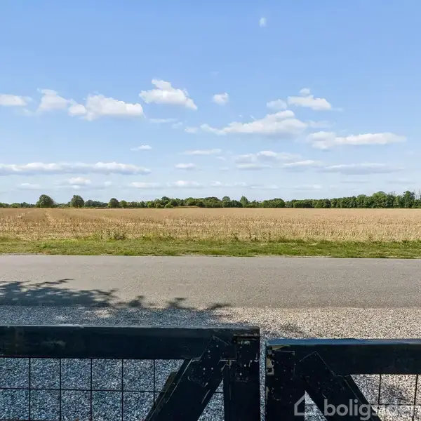 Port åbner ud mod en landevej, bag hvilken en stor mark strækker sig mod horisonten. Træer ses i baggrunden under en blå himmel med spredte skyer.