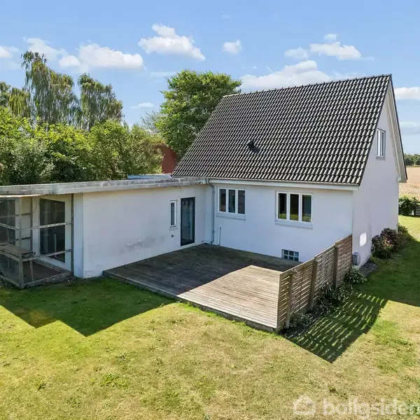 Et hvidt hus med skråtag står på en grøn græsplæne. En træterrasse foran huset fører til en større have med træer i baggrunden.