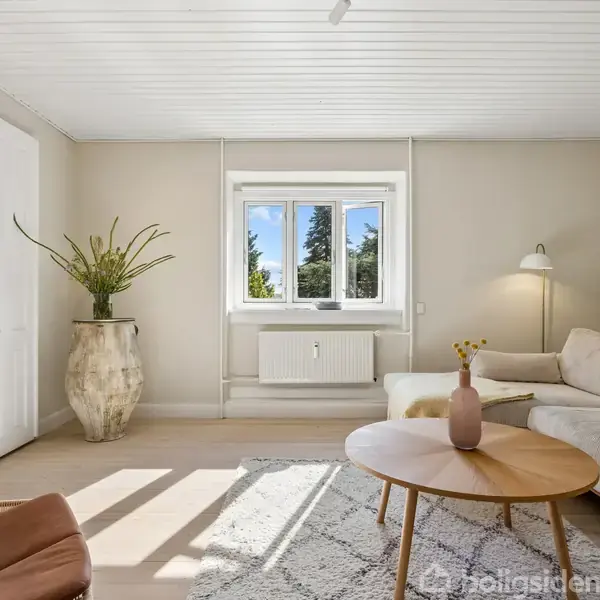 En lys stue med en beige sofa, et rundt træbord og en gulvlampe. Væggen prydes af kunst, og store vinduer viser træer udenfor. En stor vase med planter står ved døren.