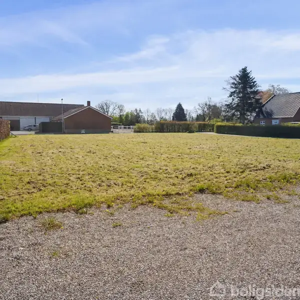 Græsplæne ligger stille i et åbent område omgivet af hække og huse under en klar blå himmel.