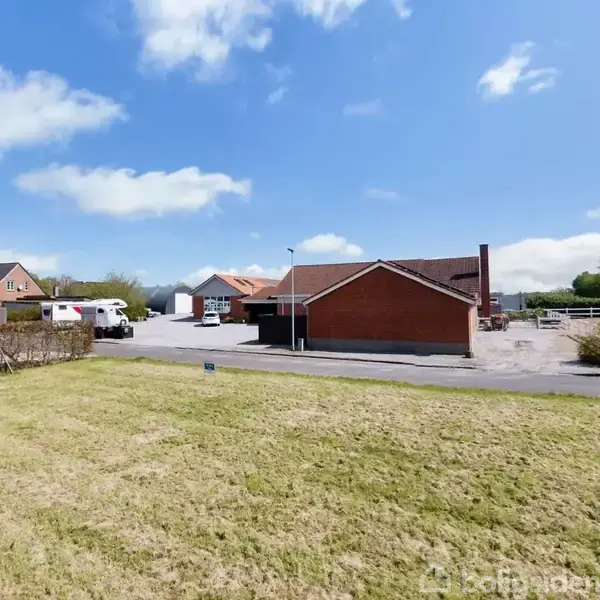 Rødt murstenshus står stille ved en vej med græsplæne i forgrunden. Biler parkerer tæt på, mens andre bygninger og en blå himmel omkranser scenen.