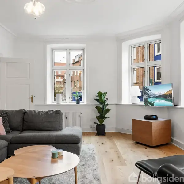 Stue med en grå sofagruppe placeret ved et rundt træbord, omgivet af lyse vægge og store vinduer. En computer står på en trækasse. Grøn plante ved vinduet. Trepunktsloftlampe i rummet.