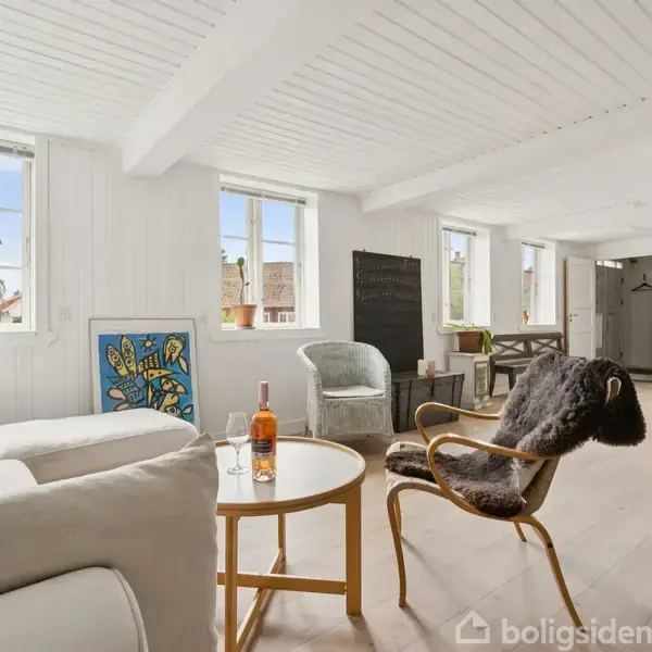 Sofa står i en lys stue med hvide vægge og loft. Et rundt bord har vinflaske og glas. Store vinduer giver udsigt, mens stole og spisebord er synlige bagved.