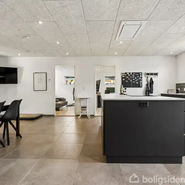 Et moderne køkken-alrum med sort køkkenø og matchende stole. Køkkenet har store fliser, hvide vægge, og indbygget belysning. En dobbeltvask er synlig ved vinduet, som har udsigt til haven.
