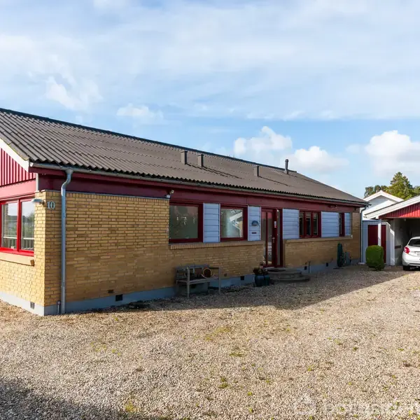 Et gult murstenshus med røde trædetaljer står stille. I baggrunden ses en carport med en bil parkeret. Området består af en grusvej og en velplejet have.