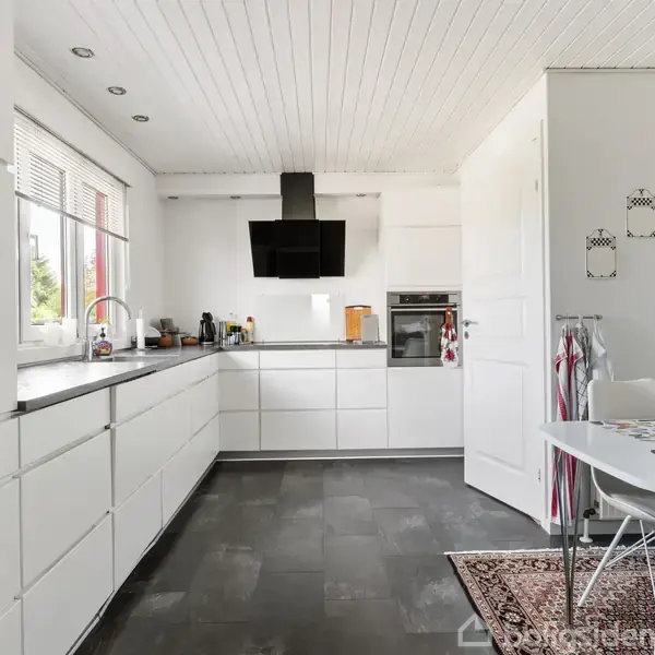 En moderne køkkenindretning med hvide skabe og integrerede apparater. Et sort emhætte hænger over komfuret. Ved siden af står et spisebord med stole på en tæppebeklædt gulv.