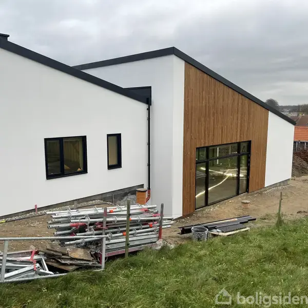 Bygning under konstruktionsfase med hvid facade og store vinduer. Rustfrit stålrør er placeret i græsset foran bygningen. Omgivet af mindre huse i baggrunden.