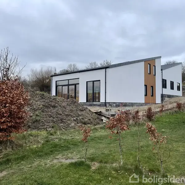 Et moderne hus står stille på en skrånende grund med nøgne træer rundt om, himlen er grå, og græsset er grønt.