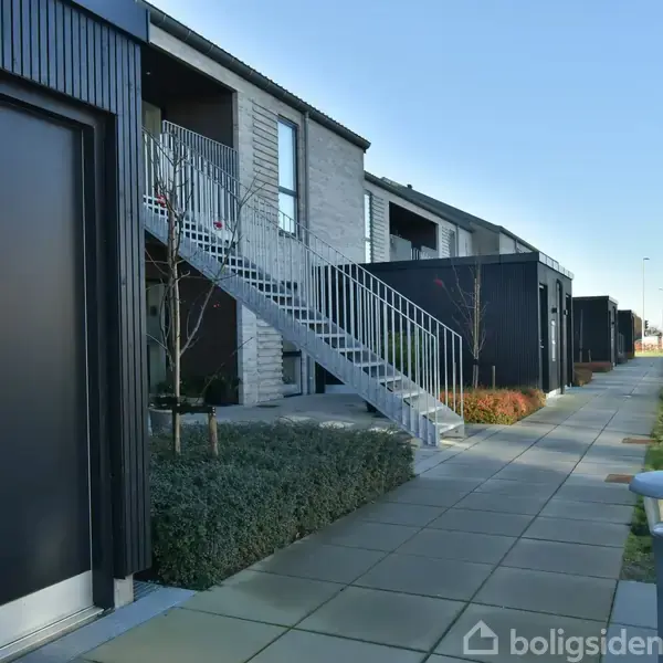 Et moderne rækkehus med mørk facade og udendørs trappe fører til en øverste etage. Omgivelserne inkluderer en flisebelagt gangsti, græsplæne og buske under en klar blå himmel.