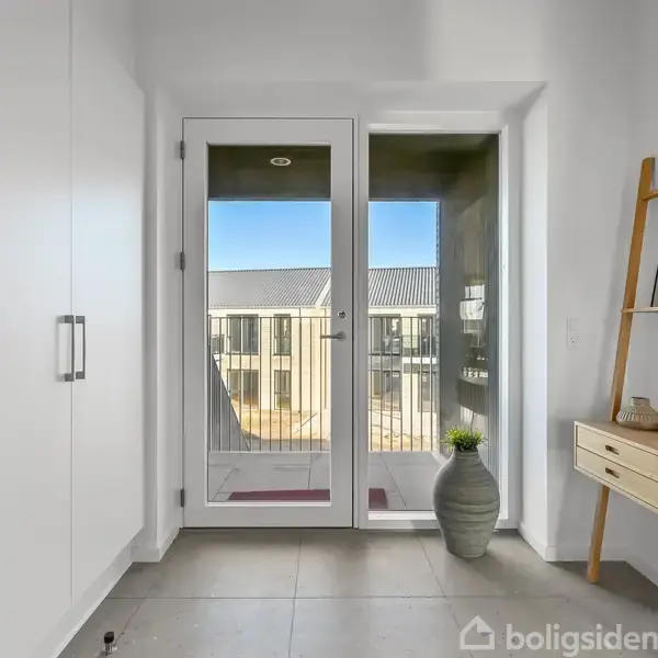 Dør med glasvindue åbner mod udendørs altan; inde er der en stor krukkeplante på gulvet, en stigehylde med dekorationer til højre og et indbygget skab til venstre.