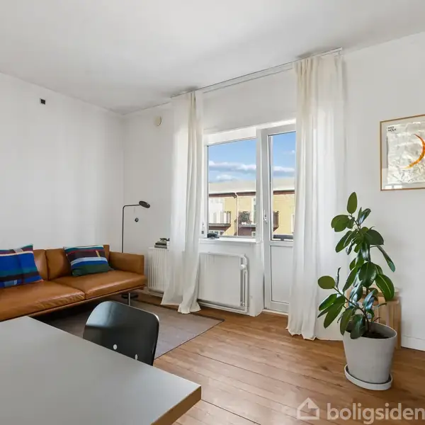 En stue indeholder en brun sofa med farverige puder. En gulvlampe står ved siden af. Store vinduer med hvide gardiner giver udsigt. En plante og hylder med bøger findes i rummet.