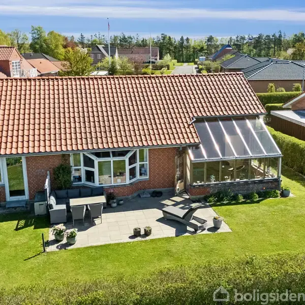 Rødt murstenshus med rødt tegltag har en terrasse med havemøbler i en velplejet have. Der er en glaskarnap til højre, omgivet af nabolagets huse.
