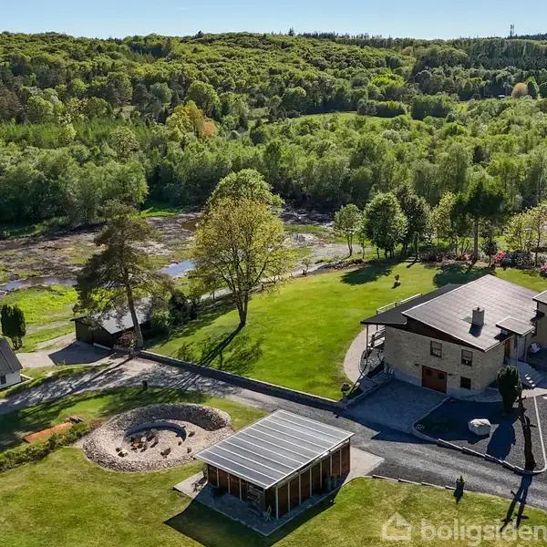 Hus står stille i frodigt landskab med grønne marker og træer. Der er en terrasse og udendørs område omkring huset, samt en lille flod i baggrunden.