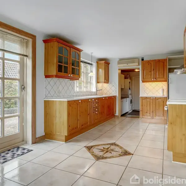 Køkken med lyse fliser, træskabe og kontrastfyldt skakternet gulvmåtte. Skabe er arrangeret langs væggen under et vindue. En dør med glasrude fører udtil en have.