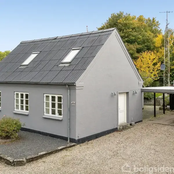 Gråt hus med mørkt tag har vinduer og hoveddør synlig. Grusbelagt gårdsplads omgivet af grønne buske og træer indrammer bygningen.