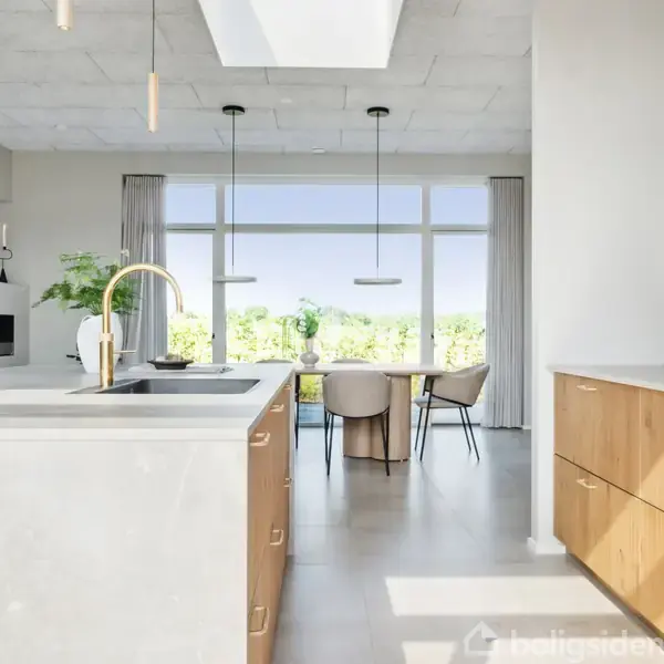 Køkkenet har en hvid marmorbordplade med en guldhane og vask. I baggrunden ses et moderne spisebord med stole og store vinduer, der giver udsigt til træer.