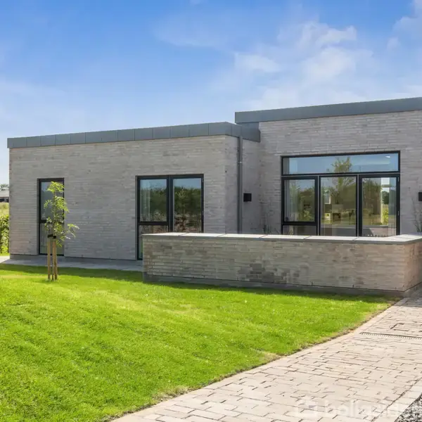 Moderne en-etagers hus med fladt tag står på en velplejet græsplæne. Store vinduer præger facaden, mens en belagt sti fører til hovedindgangen. Hækken og træerne omkranser ejendommen.