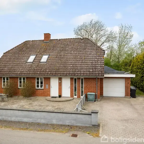 Et rødt murstenshus står stille på en stille villavej. Foran huset er der en stor indkørsel og nogle buske. Omkring huset ses grønne træer og nabohuse.