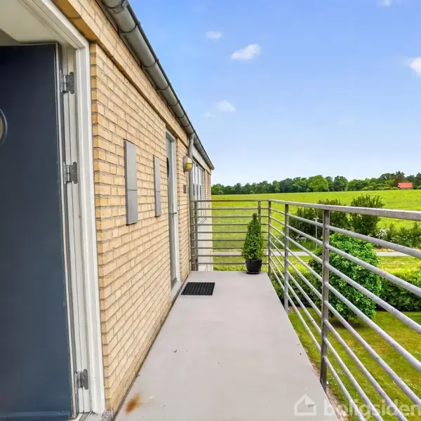 Dør står åben i en murstensbygning med metalrækværk på altan. Grønne marker og træer ses i baggrunden under en blå himmel med få skyer.