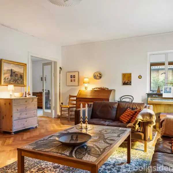 En stue med sofa og sofabord dekoreret med stearinlys. I baggrunden er der et skrivebord ved et vindue, og flere kunstværker hænger på væggene. Værelset er oplyst af lamper.