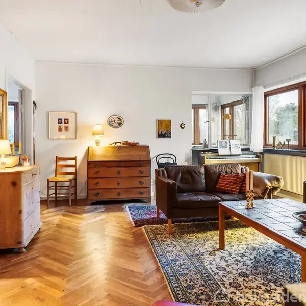 Stue med sofagruppe omkring et træbord, står på et farverigt tæppe. Reoler fyldt med bøger står langs væggen. Store vinduer lader lys komme ind, skaber en lys atmosfære.