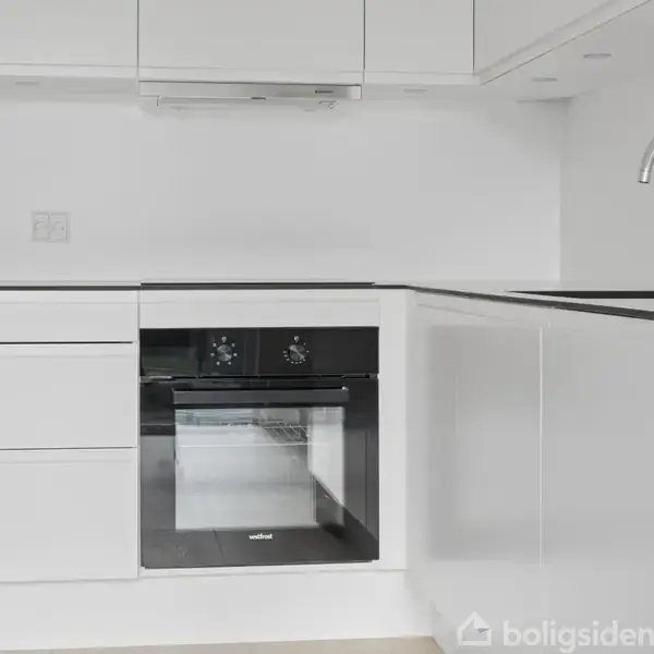 Hvidt køkken med sorte detaljer, herunder en indbygget ovn og moderne håndvask. Køkkenelementer er minimalistiske. Miljøet er rent og velorganiseret.