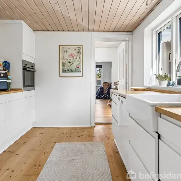 Et moderne køkken med hvide skabe og træbordplader. Der står en brødrister og en ovn på venstre side. Et tæppe ligger på det lyse trægulv, nær et vindue med planter.