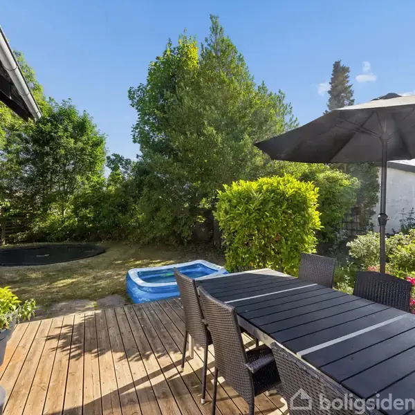 Et spisebord med stole står på en terrasse af træ. Der er en parasol over bordet. I baggrunden ses en oppustelig pool, en have og en trampolin.