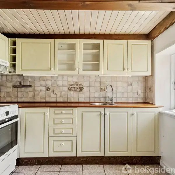 Et køkken med lysegule skabe, en indbygget ovn og kogeplade. Vasken sidder ved et vindue. Væggene er beklædt med lyse fliser, og loftet har træbjælker.