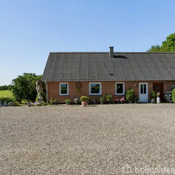 Et rødt murstenshus med gråt tag står stille foran en grusbelagt gårdsplads. Haven er prydet med planter i potter, og baggrunden viser grønne marker og træer under en blå himmel.