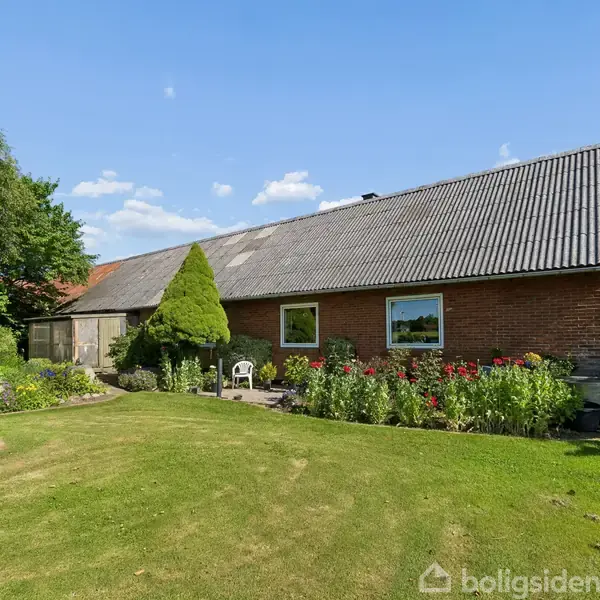 En murstensbygning med bølgeeternittag står omgivet af en velplejet have med blomsterbede og træer under en klar, blå himmel. Text: "boligsiden".