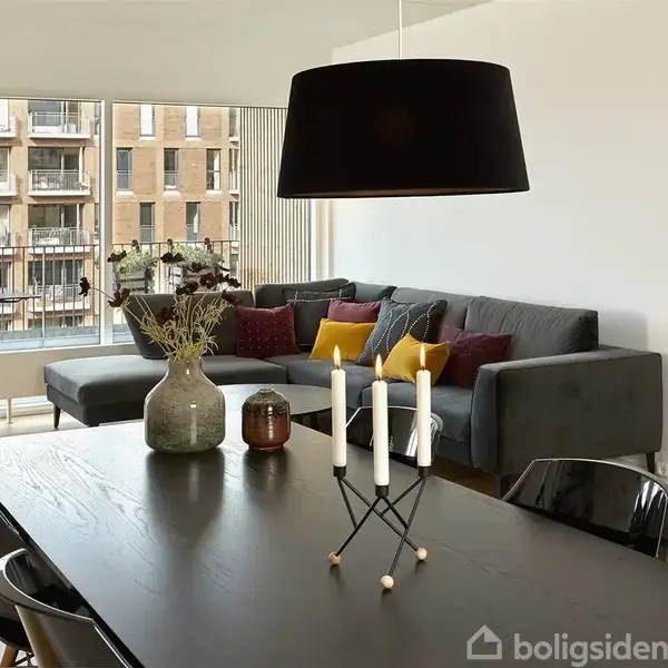 En stor, sort lampe hænger over et spisebord med stearinlys. En grå hjørnesofa med farverige puder er placeret ved store vinduer med udsigt til en bygning. Ingen tekst.