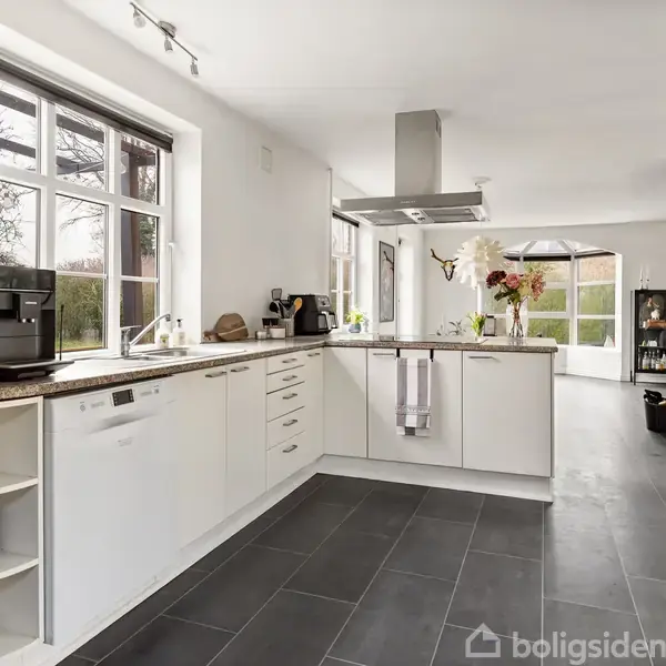Et moderne køkken med hvidt interiør og sorte fliser. Køkkenet har en kogeø med en emhætte, og der står en kaffemaskine på bordet. Stort vindue giver naturligt lys.