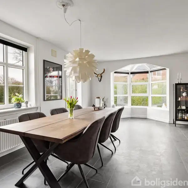 Et træbord er omgivet af seks stole i en lys spisestue med store vinduer og en moderne loftlampe. Et vitrineskab står i hjørnet nær en indbygget bænk.