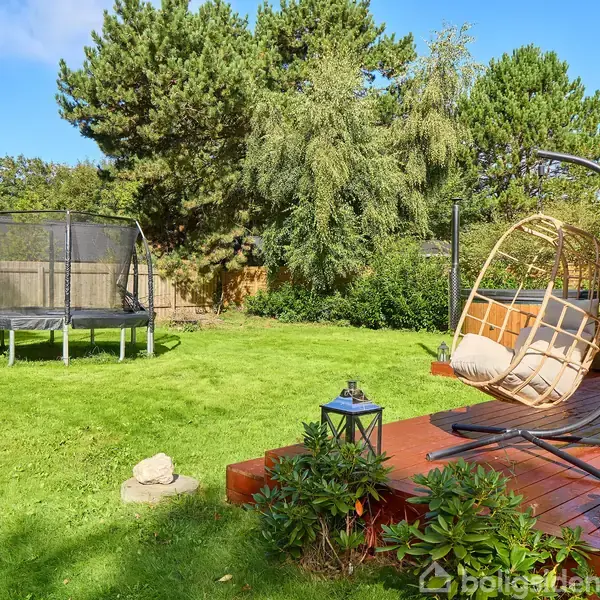Hængestol der står på en terrasse lavet af træ, omgivet af grønne buske og træer. I baggrunden er der en trampolin på en velplejet græsplæne under en blå himmel.