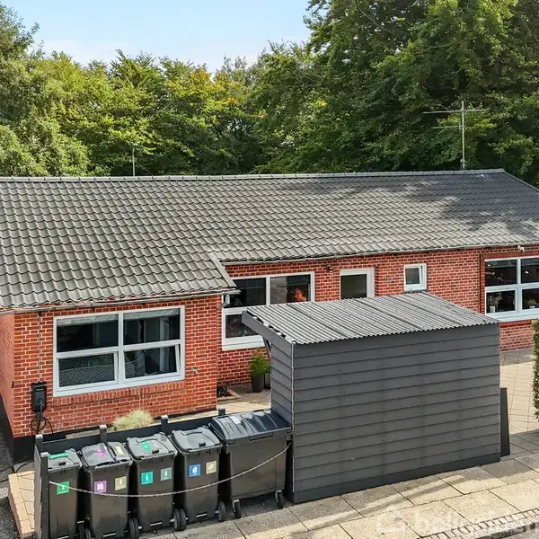 En rødmurstensbungalow med sorte skraldespande foran huset står omkranset af grønne træer. Garagen er til venstre, og en lille grå skur ses foran indkørslen.