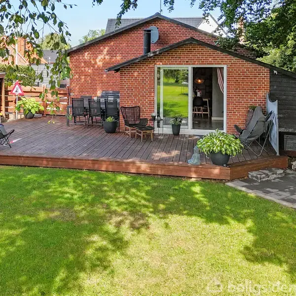 En træterrasse med havemøbler og planter foran et rødt murstenshus. Terrassen er omgivet af en grøn græsplæne med træer, og der er en åben glasdør ind til huset.