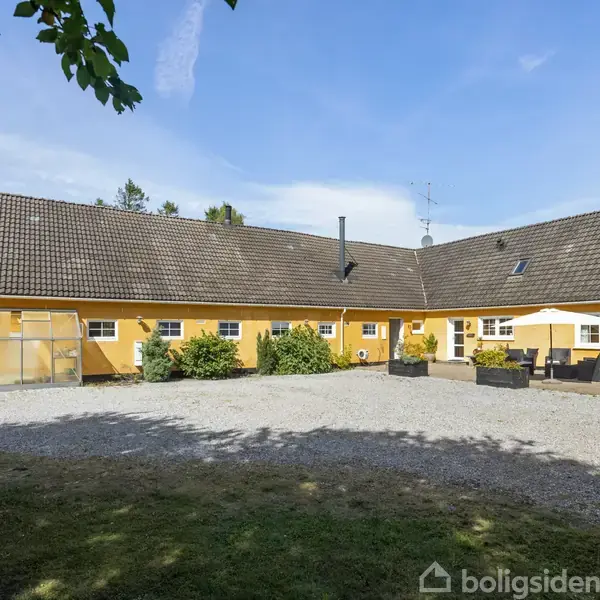Et stort, gult hus står stille med en flisebelagt gårdhave. Plantekasser og havemøbler pryder området. Omgivet af træer, under en blå himmel.