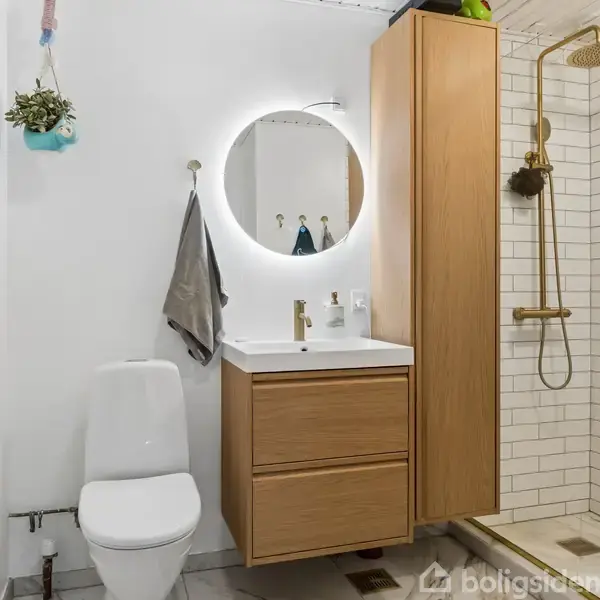 En moderne badeværelsesscene med en toilet og håndvask, der har et rundt, oplyst spejl ovenfor. Kabinen til højre har en regnbruser i en flisebelagt bruseniche.