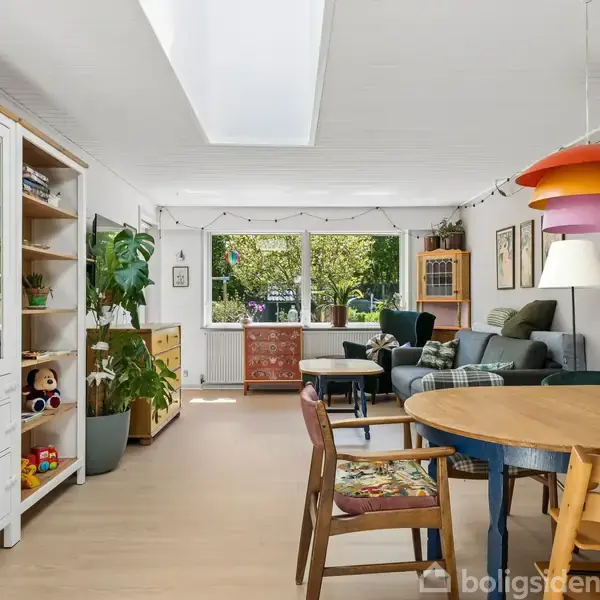 Stue med spisebord og stole; farverig lampe hænger over bordet; sofa ved vindue med udsigt til grøn have; reoler med bøger og legetøj; lys og indbydende atmosfære.