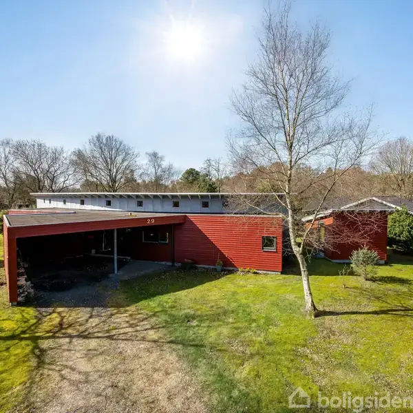 Rødt hus med fladt tag og overdækket carport står på en grøn græsplæne omgivet af træer i et åbent, solbeskinnet landskab.