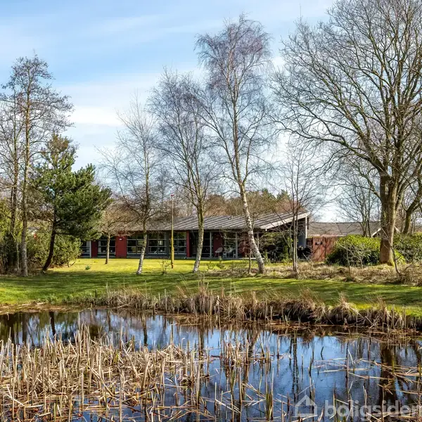 Et rødt hus står roligt blandt træer, omgivet af en naturlig have med en lille dam i forgrunden. Sollys skinner gennem det åbne landskab.