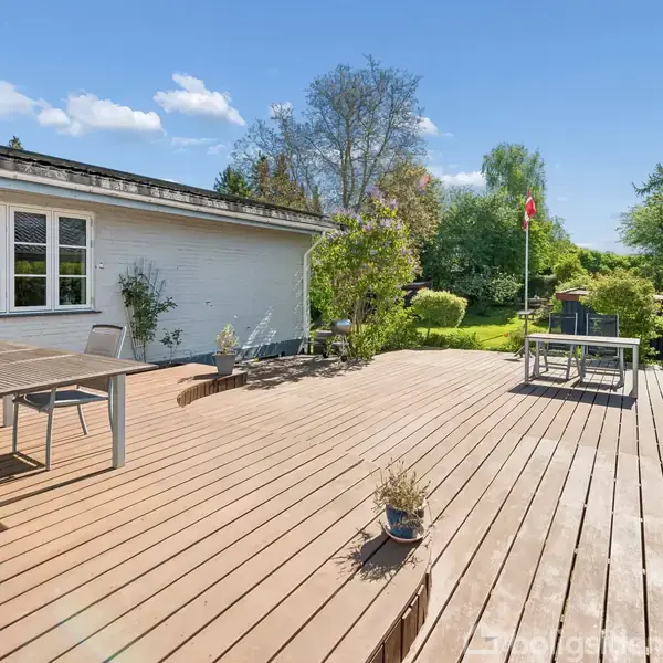 Træterrasse med bord og stole ved siden af et hus. Terrasse fører til en frodig have med træer og buske under en klar blå himmel.