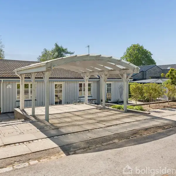 Hus med lysegrå facade og sort tag, har en åben carport foran. Omkringliggende omgivelser består af flisebelagt område og grønne buske. Klar himmel i baggrunden.
