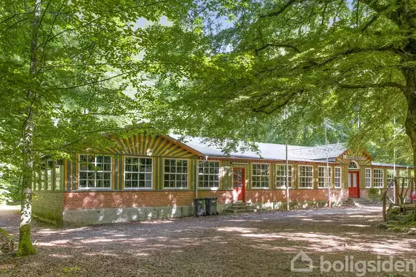 En lang, lav bygning med røde murstensvægge og mange vinduer står i en skov. Det er omgivet af grønne træer, der giver skygge og en fredfyldt atmosfære.